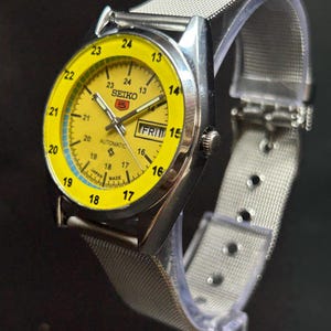 Puede incluir: Un reloj de pulsera autom&aacute;tico Seiko 5 vintage con una esfera de color amarillo brillante y una correa de malla de acero inoxidable plateada. La esfera del reloj muestra el d&iacute;a y la fecha, con una escala de tiempo de 24 horas. El reloj se muestra sobre un soporte transparente.