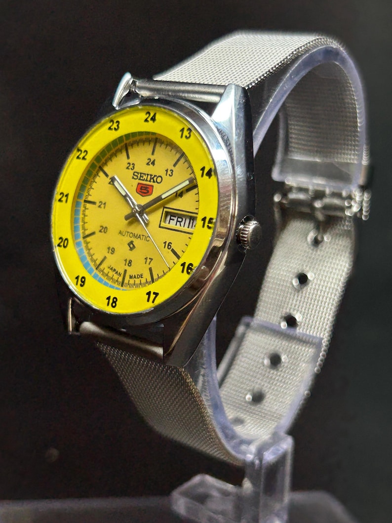 Puede incluir: Un reloj de pulsera autom&aacute;tico Seiko 5 vintage con una esfera de color amarillo brillante y una correa de malla de acero inoxidable plateada. La esfera del reloj muestra el d&iacute;a y la fecha, con una escala de tiempo de 24 horas. El reloj se muestra sobre un soporte transparente.