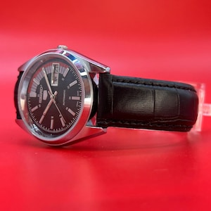 May include: A silver-toned Seiko automatic watch with a black dial and a black leather band. The watch face displays the day and date. The watch is set against a red background.
