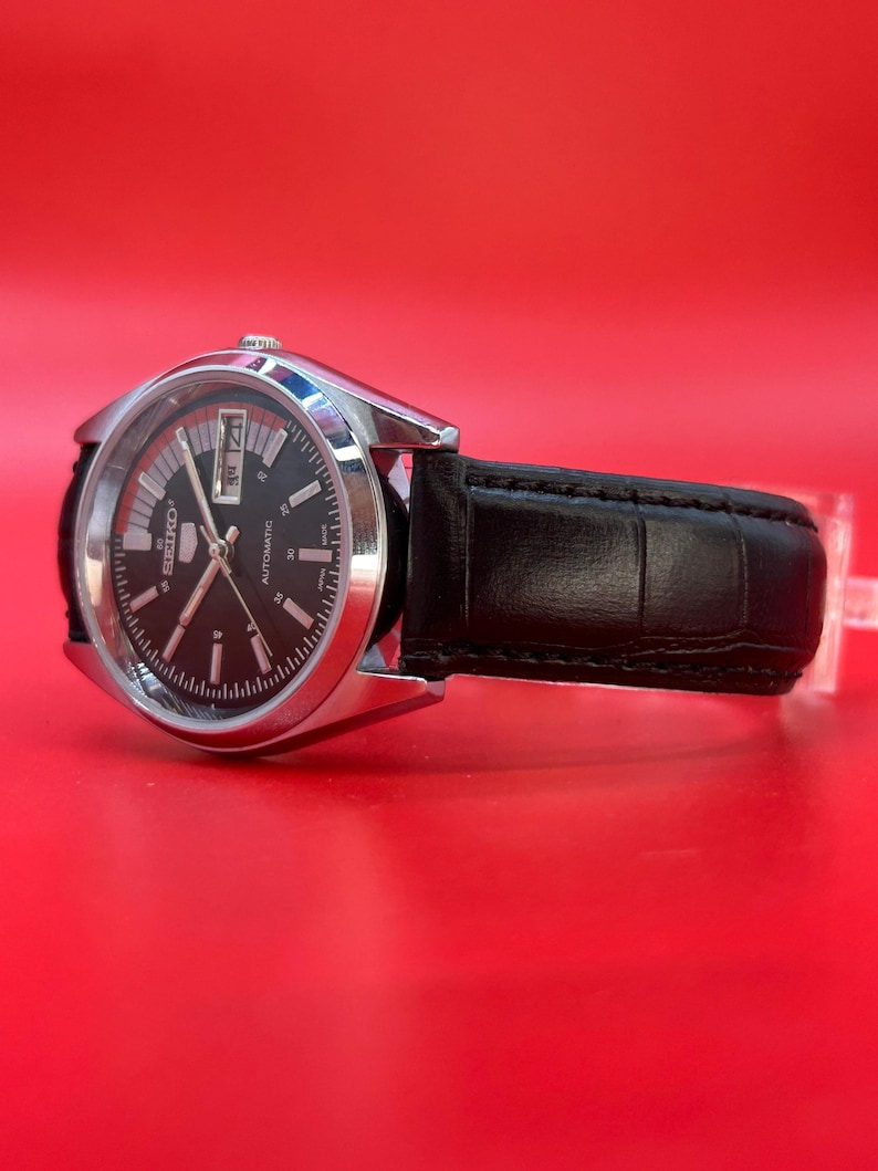 May include: A silver-toned Seiko automatic watch with a black dial and a black leather band. The watch face displays the day and date. The watch is set against a red background.