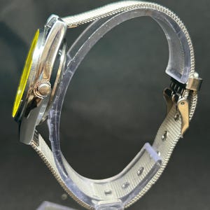 Puede incluir: Un reloj plateado con esfera amarilla y correa de malla. El reloj se muestra de lado, destacando el cierre de la correa y la esfera del reloj. El reloj est&aacute; expuesto sobre un soporte transparente.