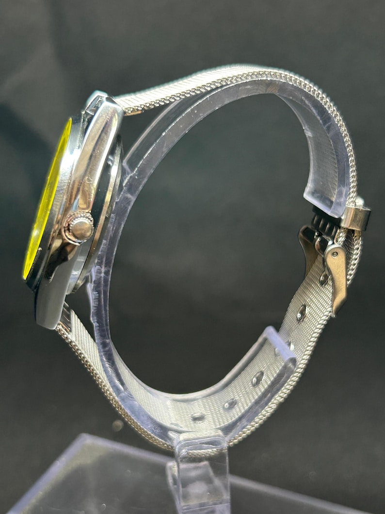 Puede incluir: Un reloj plateado con esfera amarilla y correa de malla. El reloj se muestra de lado, destacando el cierre de la correa y la esfera del reloj. El reloj est&aacute; expuesto sobre un soporte transparente.