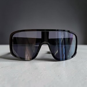 May include: Black sport sunglasses with a wraparound design. The lenses are dark, and the frames are matte black. The sunglasses have a modern, aerodynamic look, suitable for outdoor activities.