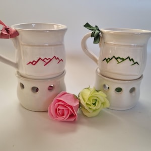 May include: Two white ceramic mugs with mountain designs, one with a red mountain and the other with a green mountain. Each mug has a handle and sits on a matching warmer. Pink and yellow roses are in front.