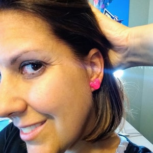 May include: A close-up of a person's ear with a pair of pink heart-shaped earrings. The earrings are small and have a sparkly finish.