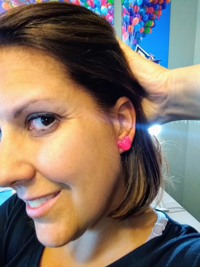 May include: A close-up of a person's ear with a pair of pink heart-shaped earrings. The earrings are small and have a sparkly finish.