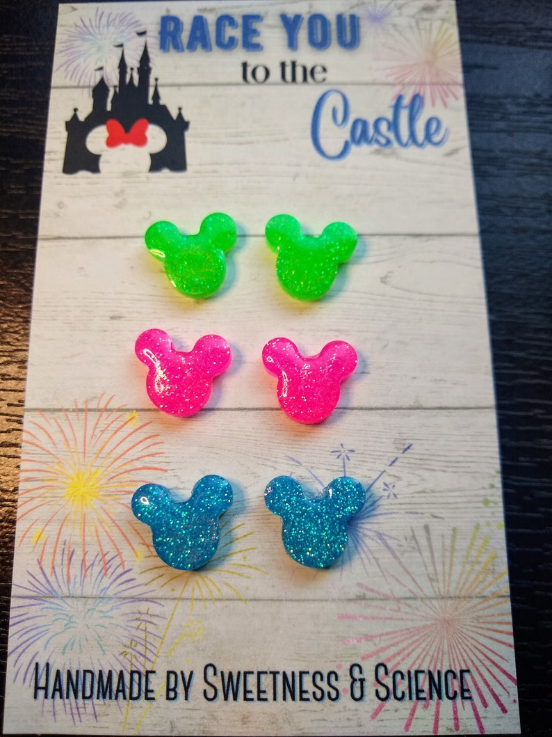 May include: A set of six glittery Mickey Mouse ear earrings in green, pink and blue. The earrings are displayed on a card with the text "Race You to the Castle" and "Handmade by Sweetness & Science".