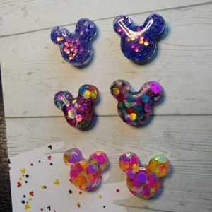 May include: Six pairs of handmade resin Mickey Mouse earrings. Each pair features a different colour combination of glitter and confetti. The earrings are displayed on a white card with the text "Handmade by Sweetness & Co."