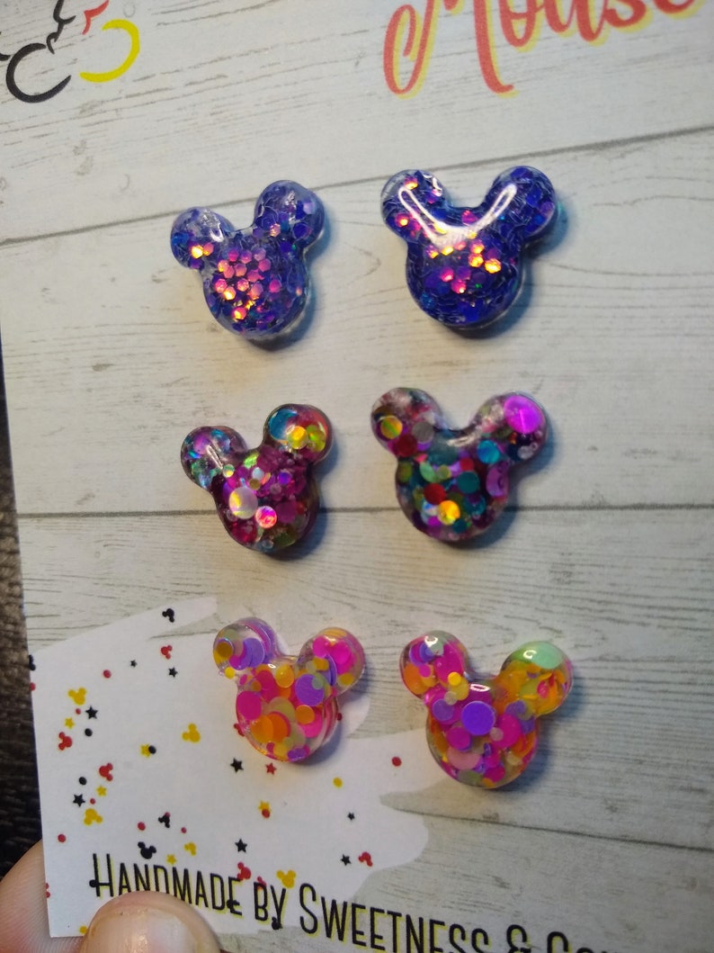 May include: Six pairs of handmade resin Mickey Mouse earrings. Each pair features a different colour combination of glitter and confetti. The earrings are displayed on a white card with the text "Handmade by Sweetness & Co."