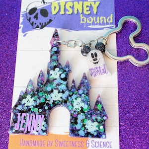 May include: A purple and green glitter resin keychain shaped like Cinderella's castle with a Mickey Mouse ghost charm. The charm says "foolish mortal". The keychain is handmade and says "Jenny" on the front. The background is a purple glitter fabric.
