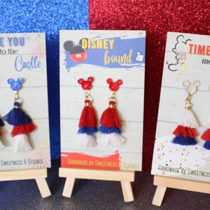May include: Three pairs of red, white, and blue tassel earrings with Mickey Mouse ears on the top. The earrings are displayed on small wooden easels with signs that say "Race You to the Castle", "Disney Bound", and "Time to Meet the Mouse".