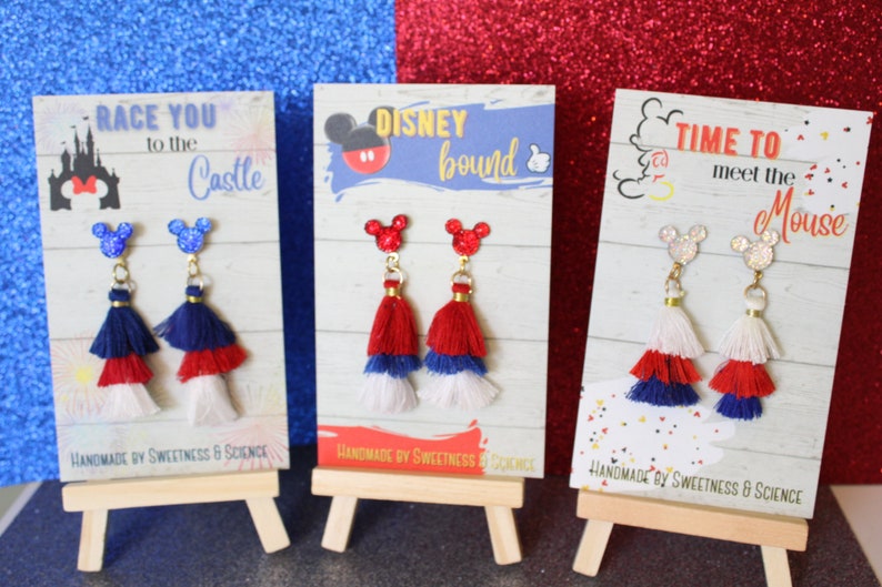 May include: Three pairs of red, white, and blue tassel earrings with Mickey Mouse ears on the top. The earrings are displayed on small wooden easels with signs that say "Race You to the Castle", "Disney Bound", and "Time to Meet the Mouse".