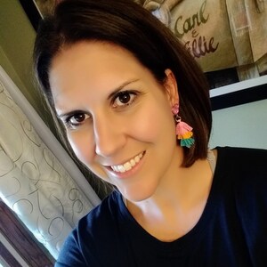 May include: A woman with brown hair wearing a black shirt and pink and green tassel earrings. The background shows a framed picture with the text "Carl & Ellie".