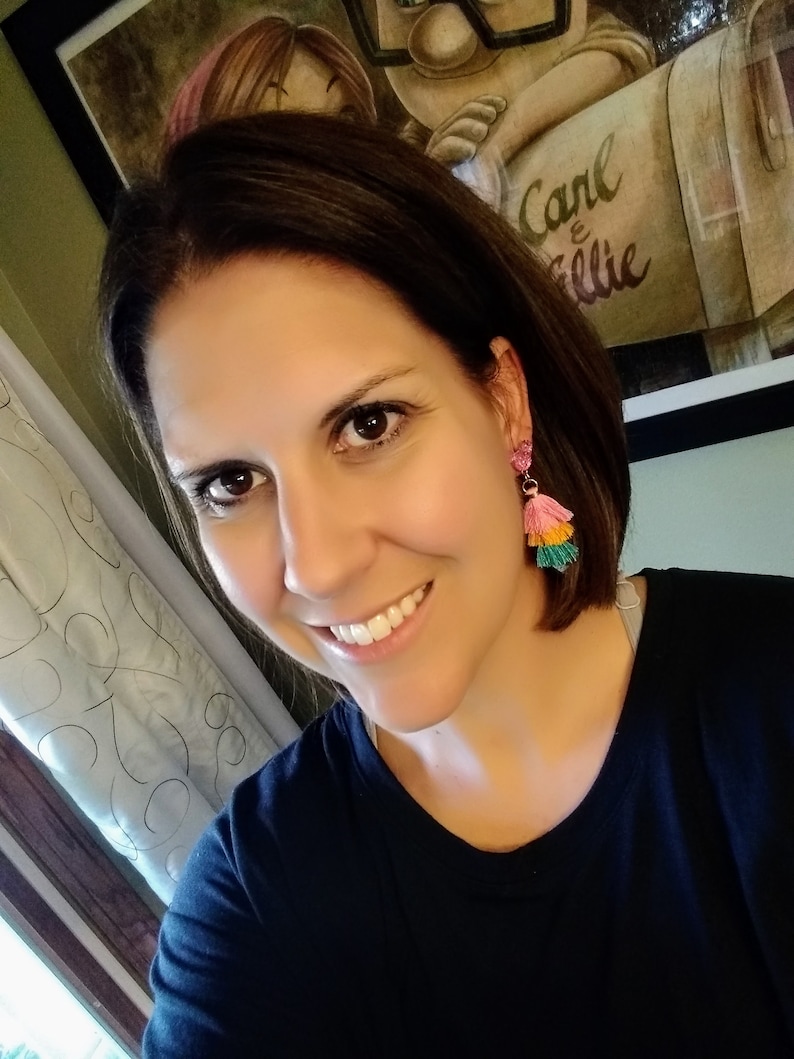 May include: A woman with brown hair wearing a black shirt and pink and green tassel earrings. The background shows a framed picture with the text "Carl & Ellie".