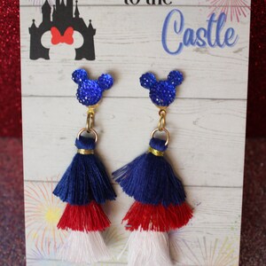 May include: A pair of blue, red, and white tassel earrings with a blue glitter Mickey Mouse charm. The earrings are displayed on a card that says "Race You to the Castle" with a silhouette of Cinderella's castle and Mickey Mouse's head with a red bow. The card also says "Handmade by Sweetness & Science".