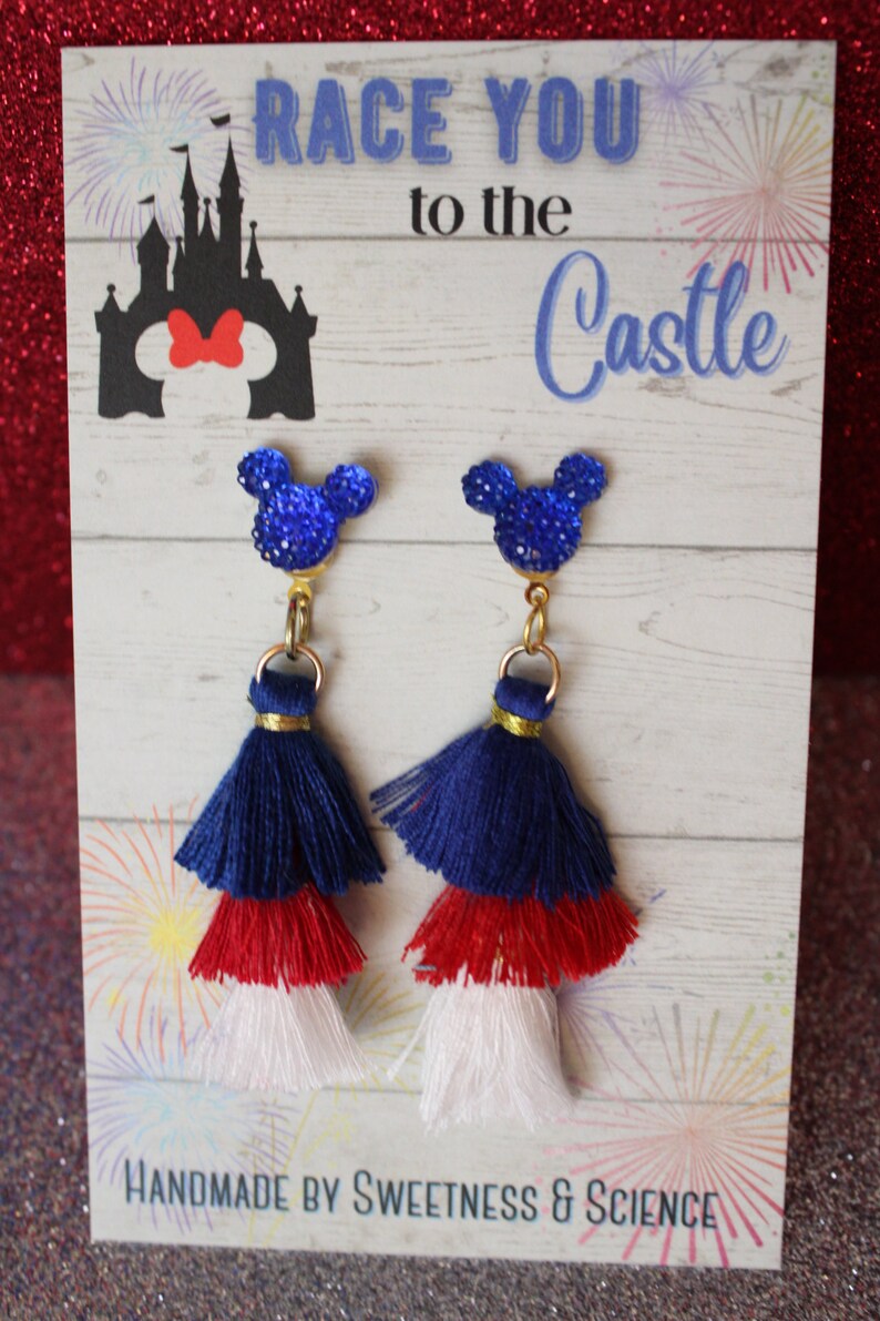 May include: A pair of blue, red, and white tassel earrings with a blue glitter Mickey Mouse charm. The earrings are displayed on a card that says "Race You to the Castle" with a silhouette of Cinderella's castle and Mickey Mouse's head with a red bow. The card also says "Handmade by Sweetness & Science".