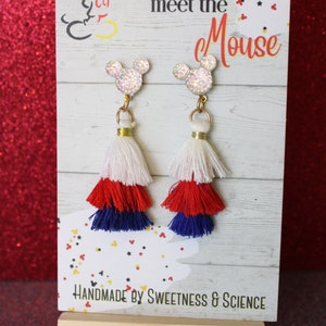 May include: A pair of red, white, and blue tassel earrings with a glitter Mickey Mouse charm. The earrings are displayed on a wooden easel with a sign that reads "Time to meet the Mouse" and "Handmade by Sweetness & Science".