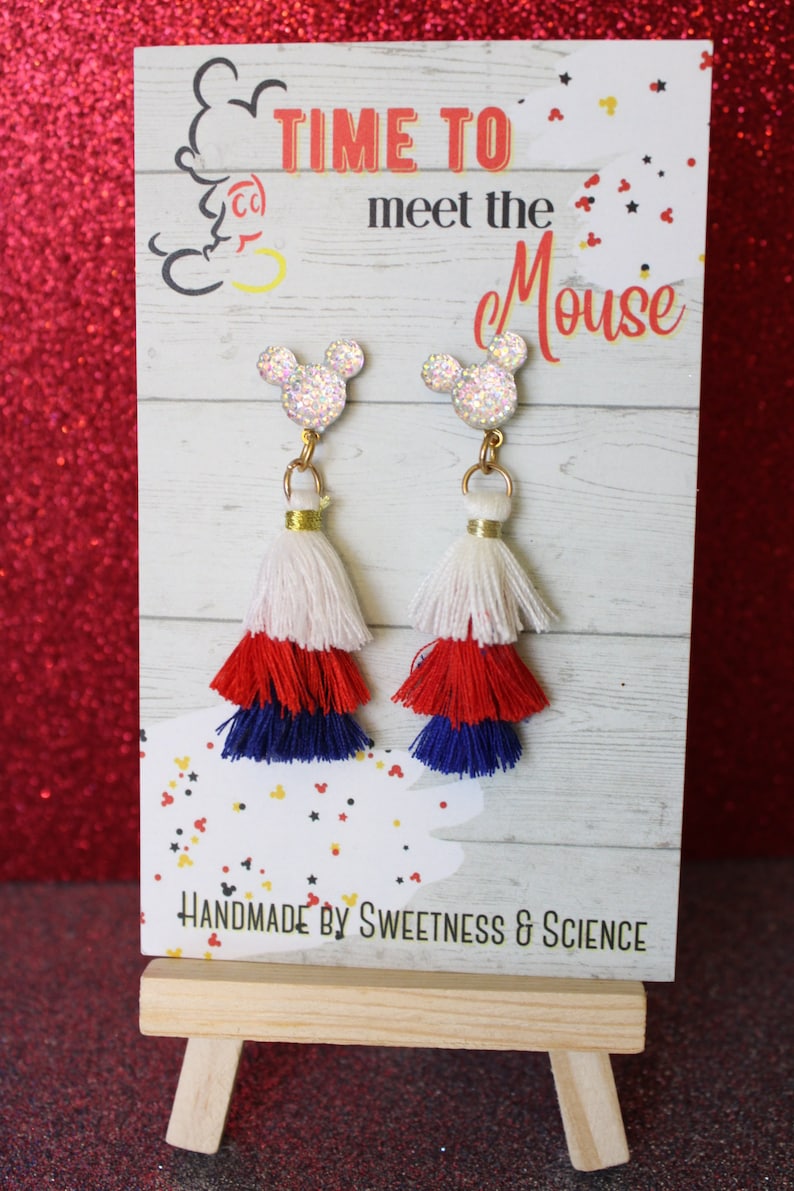May include: A pair of red, white, and blue tassel earrings with a glitter Mickey Mouse charm. The earrings are displayed on a wooden easel with a sign that reads "Time to meet the Mouse" and "Handmade by Sweetness & Science".