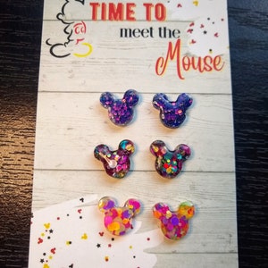 May include: A set of six colourful glitter Mickey Mouse earrings. The earrings are displayed on a card that reads "Time to meet the Mouse" and "Handmade by Sweetness & Science".