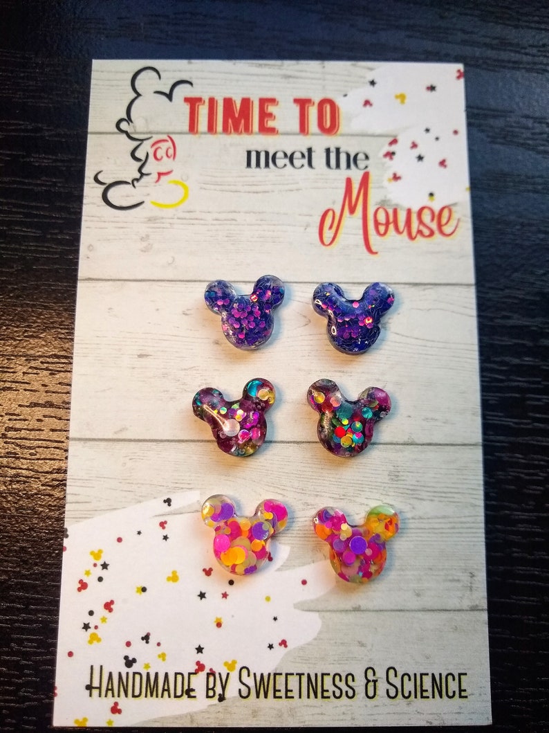 May include: A set of six colourful glitter Mickey Mouse earrings. The earrings are displayed on a card that reads "Time to meet the Mouse" and "Handmade by Sweetness & Science".