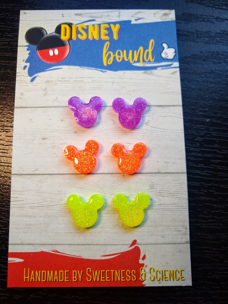 May include: Six pairs of glitter Mickey Mouse ears in purple, orange, and yellow. The ears are displayed on a white card with the text "DISNEY bound" and "HANDMADE BY SWEETNESS & SCIENCE".