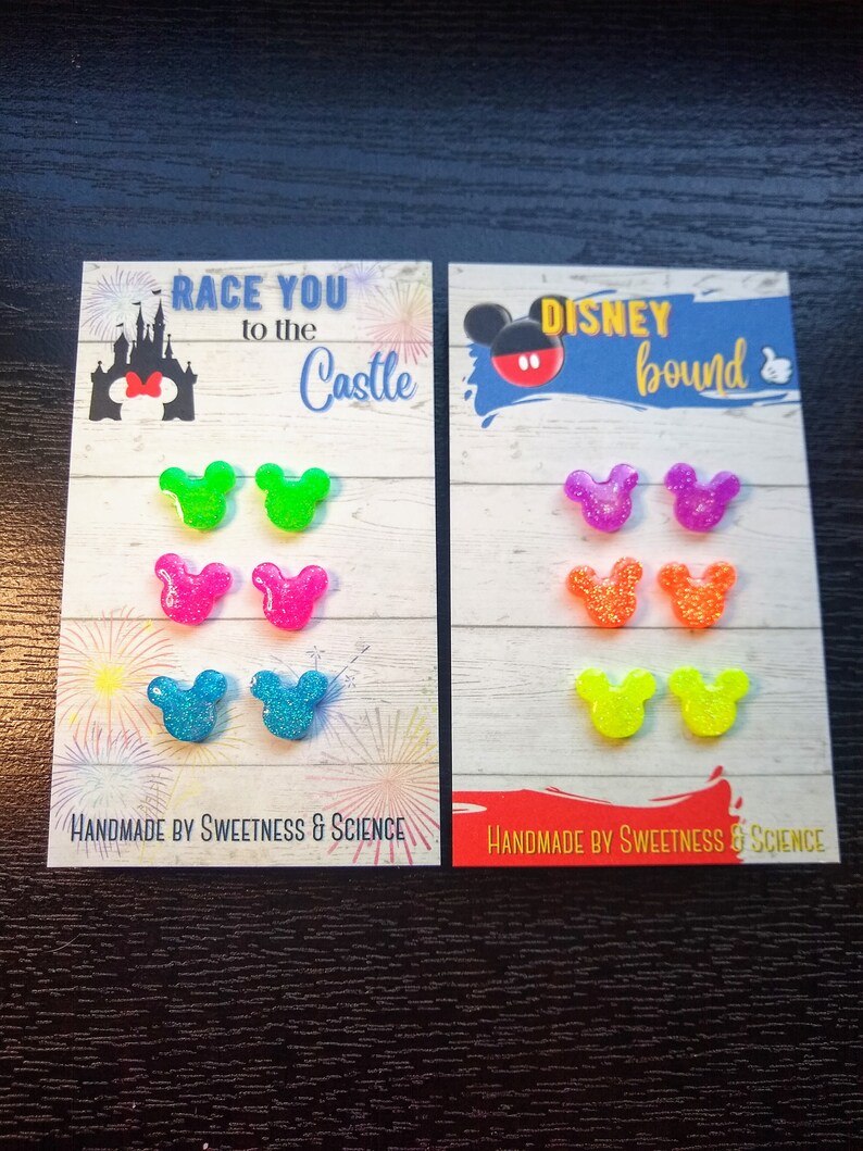 May include: Two sets of colourful Mickey Mouse ear-shaped earrings. The first set says "Race You to the Castle" and the second set says "Disney Bound". Both sets are handmade by Sweetness & Science.