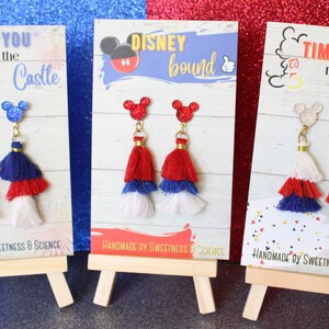 May include: Three pairs of red, white, and blue tassel earrings with Mickey Mouse head charms. The earrings are displayed on small wooden easels. The cards behind the earrings have the text "Race You to the Castle", "Disney Bound", and "Time to Meet the Mouse".