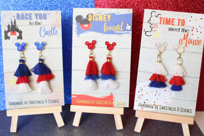 May include: Three pairs of red, white, and blue tassel earrings with Mickey Mouse head charms. The earrings are displayed on small wooden easels. The cards behind the earrings have the text "Race You to the Castle", "Disney Bound", and "Time to Meet the Mouse".