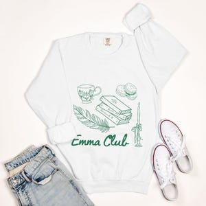 May include: White sweatshirt featuring a green illustration of a teacup, books, feather, candle, and biscuits. The words "Emma Club" are written in green script. White trainers and blue jeans are also in the image.