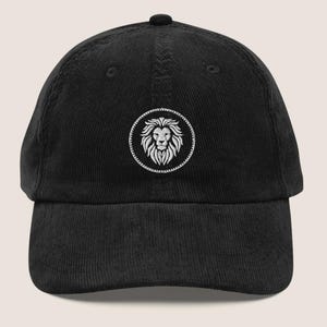 May include: Black corduroy baseball cap with a white embroidered lion crest. The cap has a curved peak and an adjustable strap. The lion crest is a detailed design within a circular border.