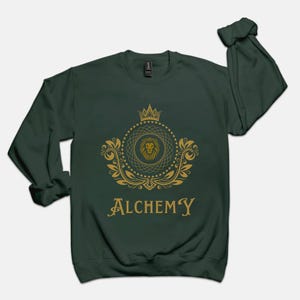 May include: Dark green sweatshirt with a gold emblem featuring a lion's head, crown, and ornate scrollwork. The word "ALCHEMY" is printed in gold script below the emblem. The sweatshirt has long sleeves and a crew neck.