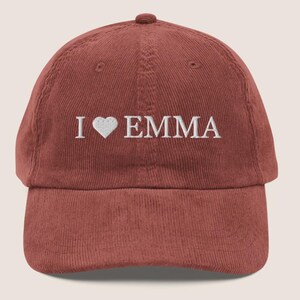 May include: A corduroy baseball cap in a reddish-brown hue. The cap features the embroidered text "I ♥ EMMA" in white, with a heart symbol replacing the word "love". The hat has a curved brim and an adjustable strap.