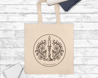 Puede incluir: Bolso tote de color crema con un diseño circular que presenta una vela en un candelabro, rodeada de elementos florales. El texto "ON TRAITORS ROAD" está impreso debajo. El bolso tiene dos asas y está hecho de un material suave.