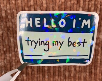 以下が含まれることがあります： 上部に「HELLO I'M」、下部に「trying my best」と書かれた長方形のステッカー。ステッカーは、ホログラフィックで虹色のボーダーが付いたティール色の背景です。ピンセットがステッカーを持っています。