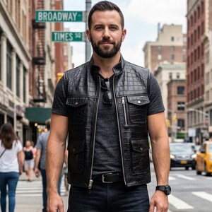 Op de afbeelding: Een persoon draagt een zwart leren vest met een gewatteerd patroon, een grijs t-shirt, donkerblauwe jeans en zwarte laarzen. Ze staan op een stoep in de stad met gebouwen en verkeer op de achtergrond. Een zwart horloge zit om hun linkerpols.