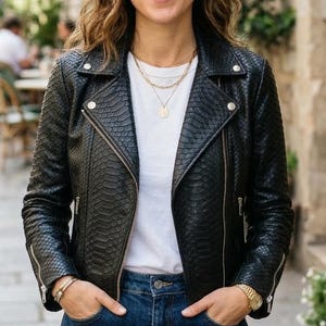 May include: A person wearing a black leather jacket with a snakeskin pattern, a white t-shirt, and blue jeans. She is standing on a cobbled street, smiling. The jacket has a zip closure and silver hardware. The background shows a European street.