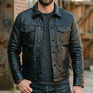 May include: A black leather jacket with a snakeskin effect. The jacket features two chest pockets, button fastenings, and a collar. The person is wearing a grey t-shirt and dark wash jeans.