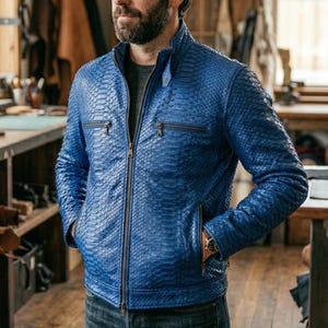 May include: A vibrant blue leather jacket with a textured snakeskin pattern. The jacket features a zip-up front, two chest pockets, and a stand-up collar. The person is wearing a watch. The jacket is a stylish piece of outerwear.
