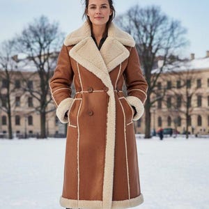 May include: A long brown coat with a cream-colored shearling lining and trim. The coat features a double-breasted design, a wide collar, and a belt at the waist. The model is wearing black pants and brown boots. The background is a snowy outdoor scene.