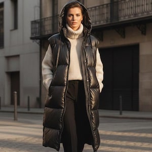 May include: A woman wearing a long black puffer vest with a hood, over a cream-colored turtleneck sweater and black pants. The vest is knee-length and has a quilted design. She is wearing black boots and walking on a cobblestone street.