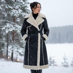 May include: A long black coat with a cream-colored shearling lining and trim. The coat features a belted waist, button closures, and a classic lapel collar. The person is wearing black boots and standing in a snowy forest.