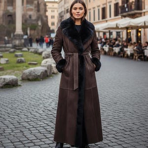May include: A long, dark brown coat featuring a black faux fur collar and black faux fur cuffs. The coat is floor-length, includes a matching fabric belt tied at the waist, and has a structured, paneled design that extends down to the hemline.