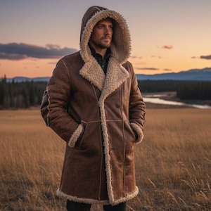 May include: A brown hooded coat with a cream-coloured shearling lining and trim. The coat has a button closure and pockets. The person is standing in a field of tall grass with a river and mountains in the background.