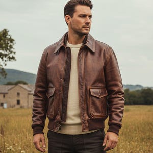 Könnte beinhalten: Braune Leder-Bomberjacke mit Kragen, zwei Vordertaschen und geripptem Saum und Bündchen. Die Jacke wird über einem cremefarbenen Pullover getragen. Der Hintergrund zeigt ein Feld und ein Gebäude.