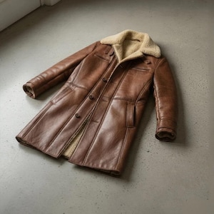 K&ouml;nnte beinhalten: Ein brauner Ledermantel mit einem Shearling-Kragen und -Futter. Der Mantel hat eine Knopfleiste, zwei Vordertaschen und ein klassisches, langes Design. Der Mantel ist mittelbraun und scheint aus hochwertigem Leder gefertigt zu sein.