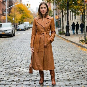 May include: A long, tan leather trench coat with a matching belt and knee-high boots. The coat is worn by a person standing on a cobblestone street. The coat has a classic trench coat design with a collar and pockets. The person is also holding a small brown purse.