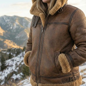May include: Brown leather jacket with a shearling collar and lining. The jacket features a zip-up front, pockets, and buckle details. The jacket is worn outdoors with a mountain backdrop.