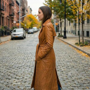 May include: A long, brown leather trench coat with a belted waist and side pockets. The coat is knee-length and has a classic, tailored design. The model is standing on a cobblestone street.