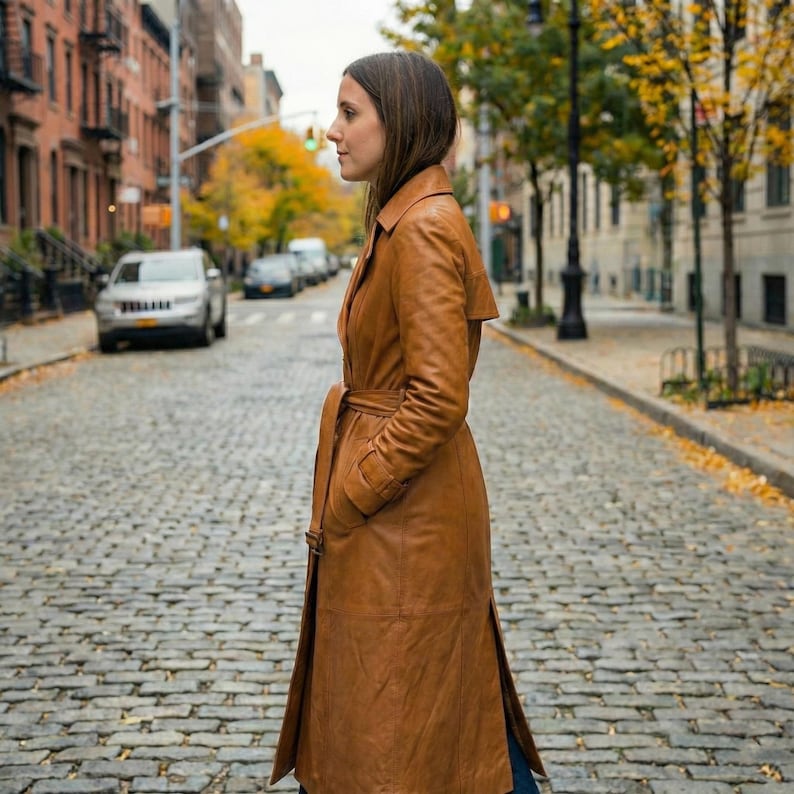 May include: A long, brown leather trench coat with a belted waist and side pockets. The coat is knee-length and has a classic, tailored design. The model is standing on a cobblestone street.