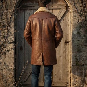 K&ouml;nnte beinhalten: Ein brauner Ledermantel mit cremefarbenem Shearling-Kragen. Der Mantel ist knielang und hat ein klassisches Design. Die Person tr&auml;gt eine blaue Jeans. Im Hintergrund eine Holzt&uuml;r und eine Steinmauer.